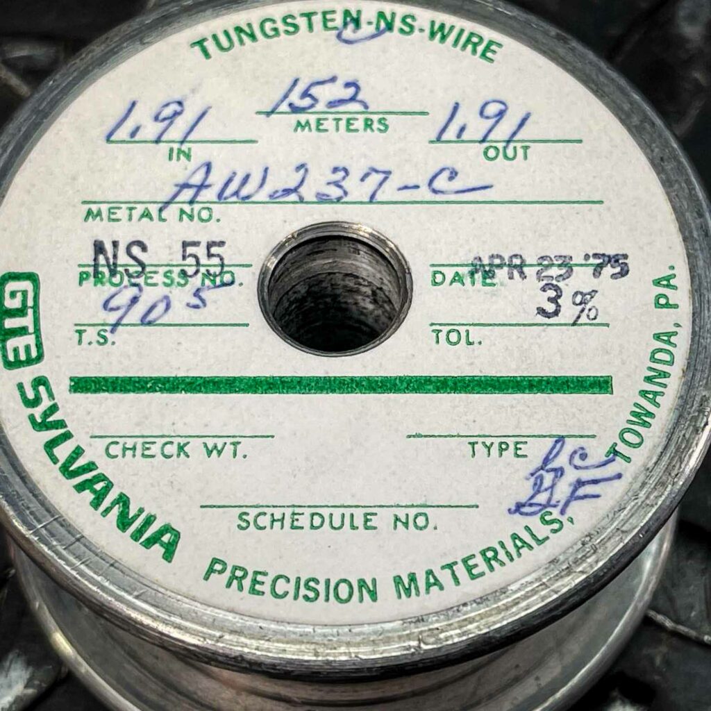 A roll of tungsten wire from 1975 for sale from Obtainium Science and Surplus. The image focuses on the top of the spool, showing a white label with green and blue handwritten text, including specifications like "NS 55," "AW237-C," and "1.91 METERS." The label is slightly worn but still legible. The spool has a central hole and rests on a textured metal surface.