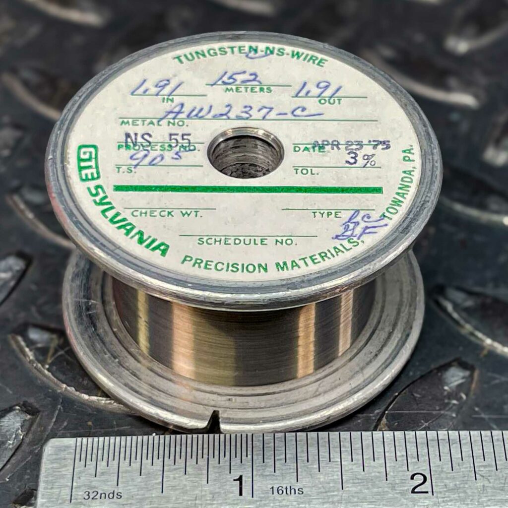 A roll of tungsten wire from 1975 for sale from Obtainium Science and Surplus. The spool is metallic with a white label on top displaying handwritten and printed details, including "TUNGSTEN-NS-WIRE," "GTE SYLVANIA," and "APR 23 '75." The wire is neatly wound around the spool, which rests on a textured metal surface. A ruler at the bottom of the image provides scale.