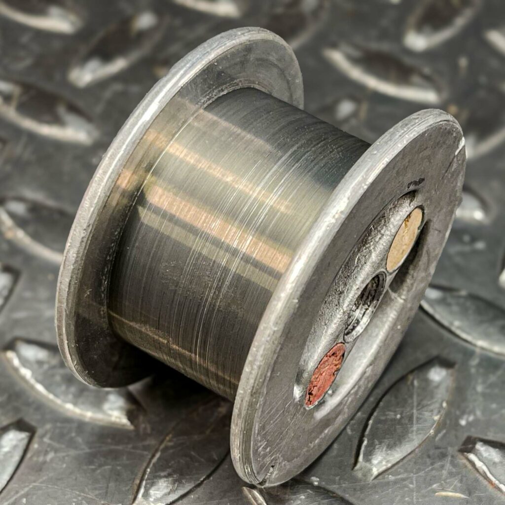 A roll of tungsten wire from 1975 for sale from Obtainium Science and Surplus. The image shows the spool from the side, revealing a darkened, metallic wire wound tightly around it. The spool appears aged, with signs of wear and oxidation. The background is a textured metal surface.