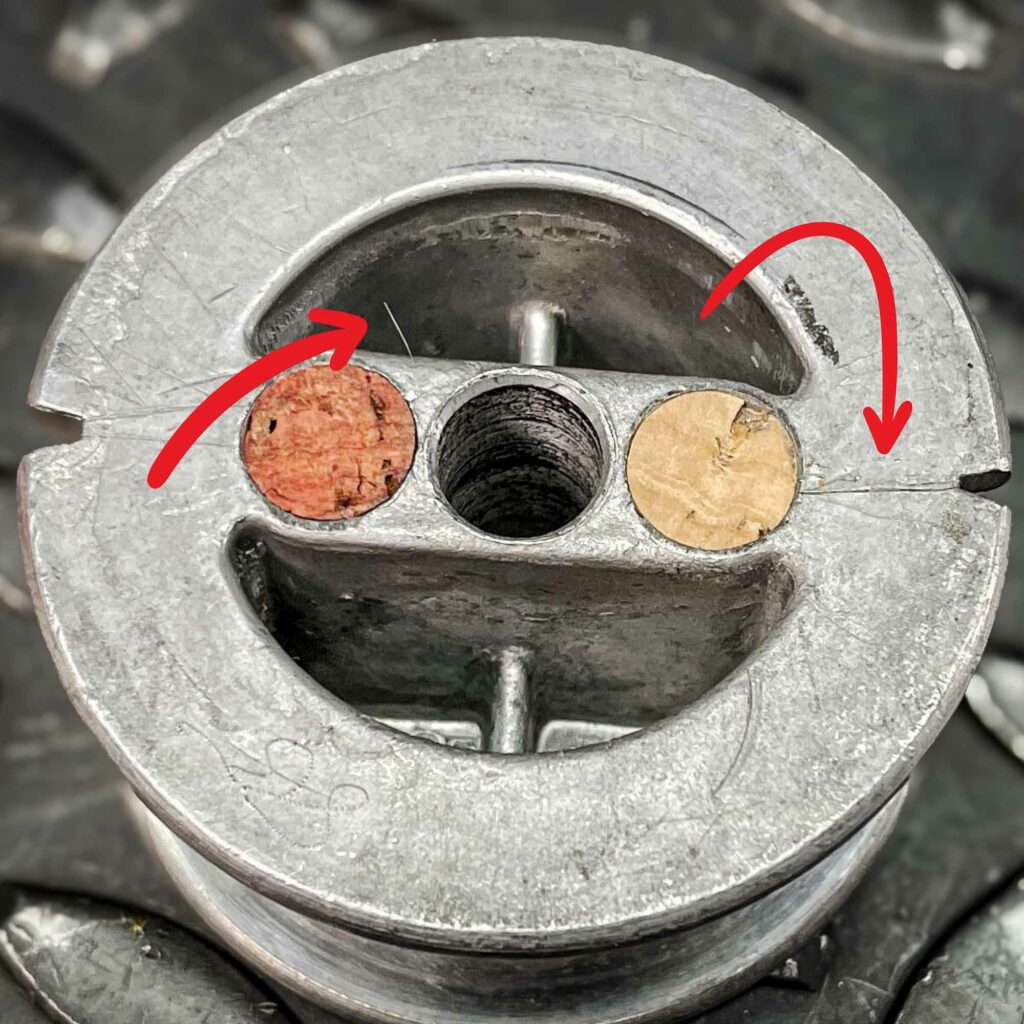 Close-up of the bottom of a roll of tungsten wire from 1975 for sale from Obtainium Science and Surplus, highlighting cracks in the metal spool. Two red arrows indicate the damaged areas. The spool's base has circular cutouts, with some filled with cork-like material. The surface appears aged and oxidized.