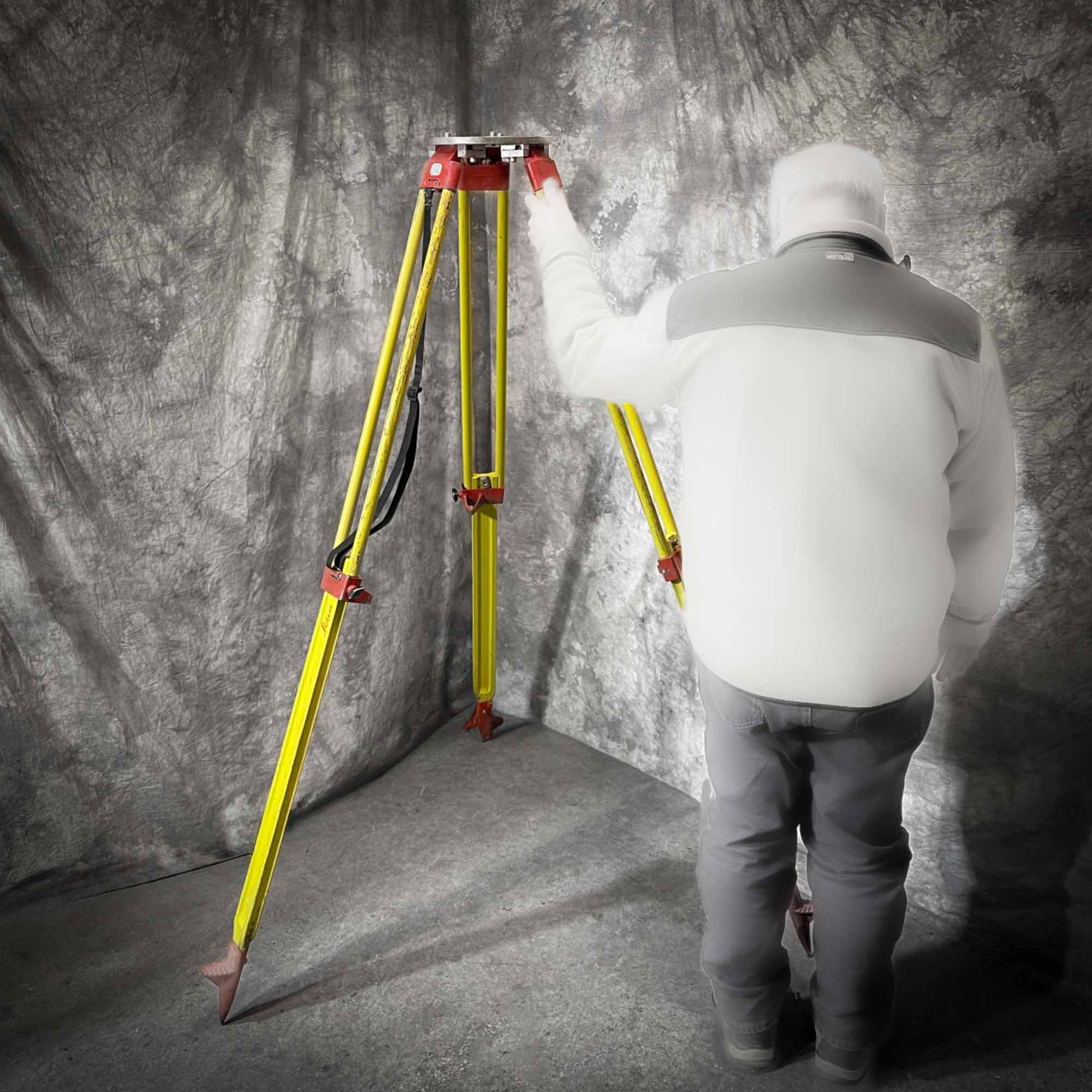 A person wearing a white jacket and dark pants stands next to a Leica wood tripod from Obtainium Science and Surplus, adjusting one of its yellow legs. The tripod has red connectors and is set up against a mottled gray backdrop.