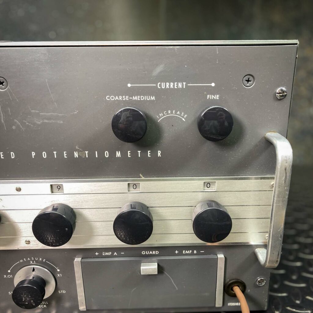 A partial close-up of the Leeds & Northrup Model 7556 Guarded Potentiometer from Obtainium Science and Surplus, focusing on the right side. Several control knobs and a handle on the metal casing are visible. The labels for voltage adjustments and settings are printed on the front panel.