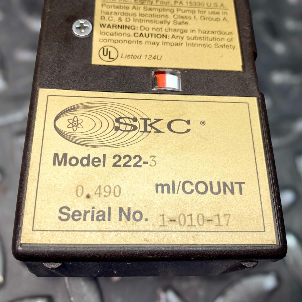 A close-up of the SKC low-flow personal air sampler model 222 from Obtainium Science and Surplus, showing the yellow label with the SKC logo, model number, and serial details. The textured metal background contrasts with the device's dark brown casing.