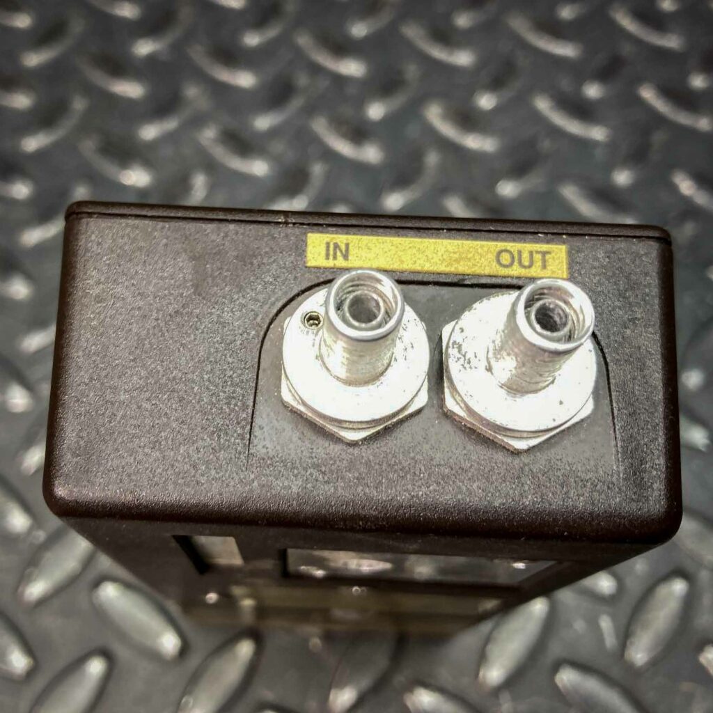 A top-down close-up of the SKC low-flow personal air sampler model 222 from Obtainium Science and Surplus, highlighting two metal air inlet and outlet ports labeled "IN" and "OUT" on a yellow strip. The textured metal background provides contrast.