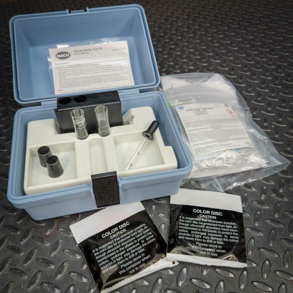 Hach Water Test Kit for Nitrites and Nitrates from Obtainium Science and Surplus, displayed open on a textured metal surface. The blue plastic case contains glass test tubes, black caps, a dropper, and compartments for reagents. Instruction sheets and reagent packets are visible.