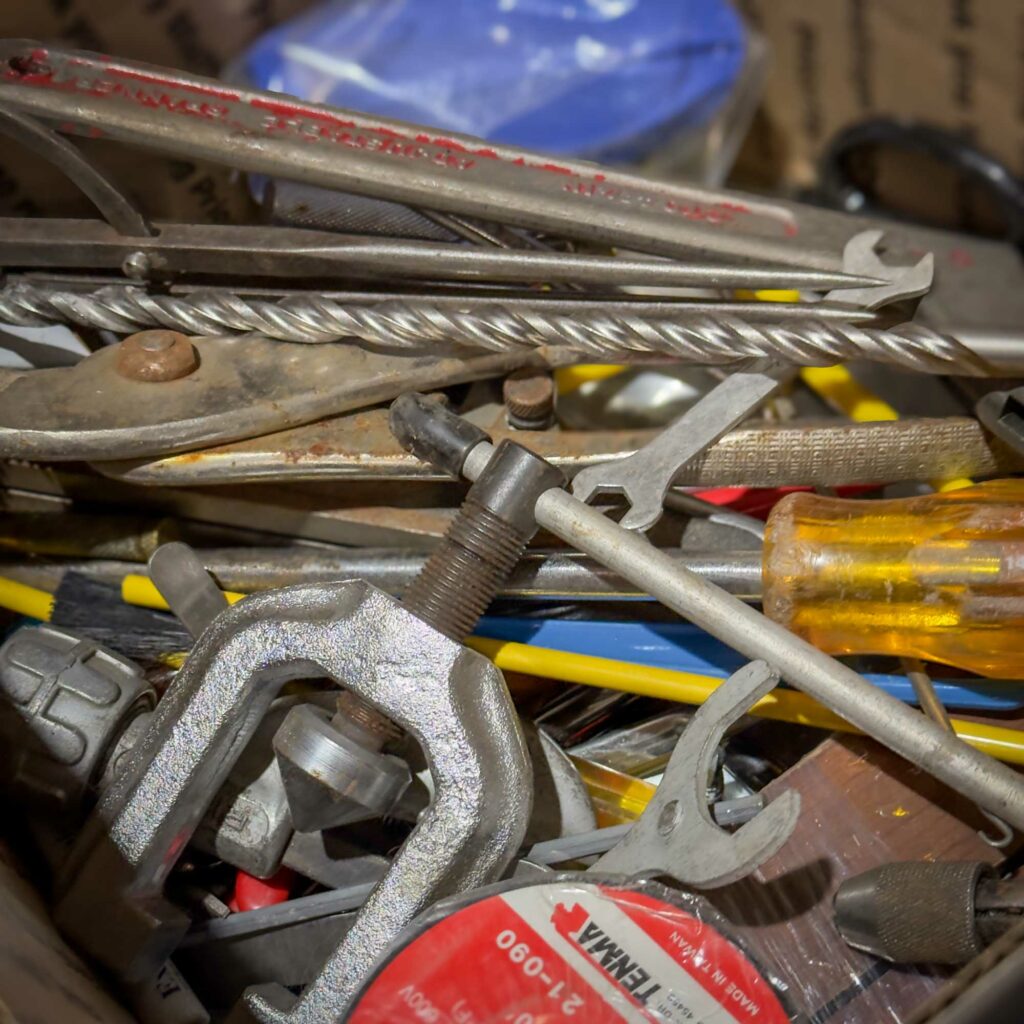 A close-up of a chaotic pile of hand tools, including wrenches, pliers, a screwdriver with a translucent yellow handle, a metal clamp, and a spool of red tape. A thick metal cable and other hardware components are also visible. A sample of what MIGHT be in the Mystery Box you get from Obtainium Science and Surplus.