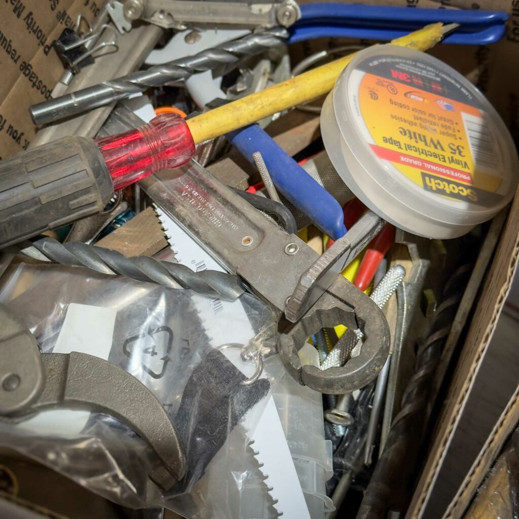 A close-up of a box filled with assorted tools and hardware, including drill bits, wrenches, screwdrivers, electrical tape, and a saw blade keychain in a plastic bag. The tools are piled chaotically, with various colors and materials visible. A sample of what could POSSIBLY be in the Mystery Box you get from Obtainium Science and Surplus.