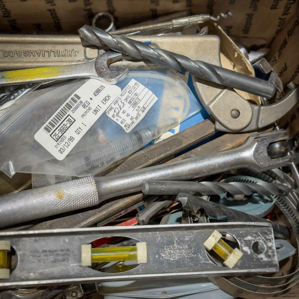 A close-up of a box filled with various tools and hardware, including drill bits, a wrench, a metal level, a syringe in a plastic bag with a label, and other miscellaneous metal components. The tools are piled in a random assortment. A sample of what could POTENTIALLY be in the Mystery Box you get from Obtainium Science and Surplus.