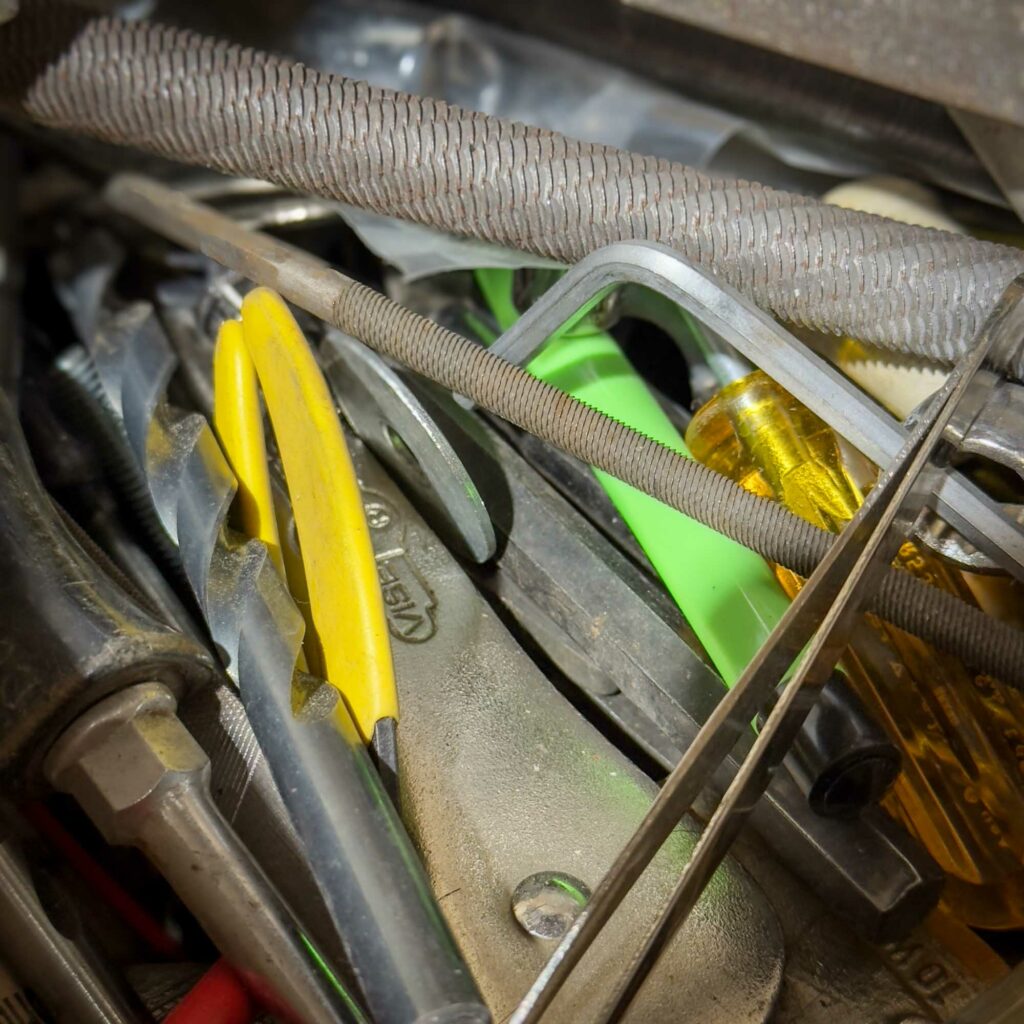 A close-up of a pile of assorted hand tools, including pliers, screwdrivers, metal files, and hex keys, in a cluttered arrangement. Some tools have yellow, green, and translucent handles, while others are metallic. A sample of what COULD be in the Mystery Box you get from Obtainium Science and Surplus